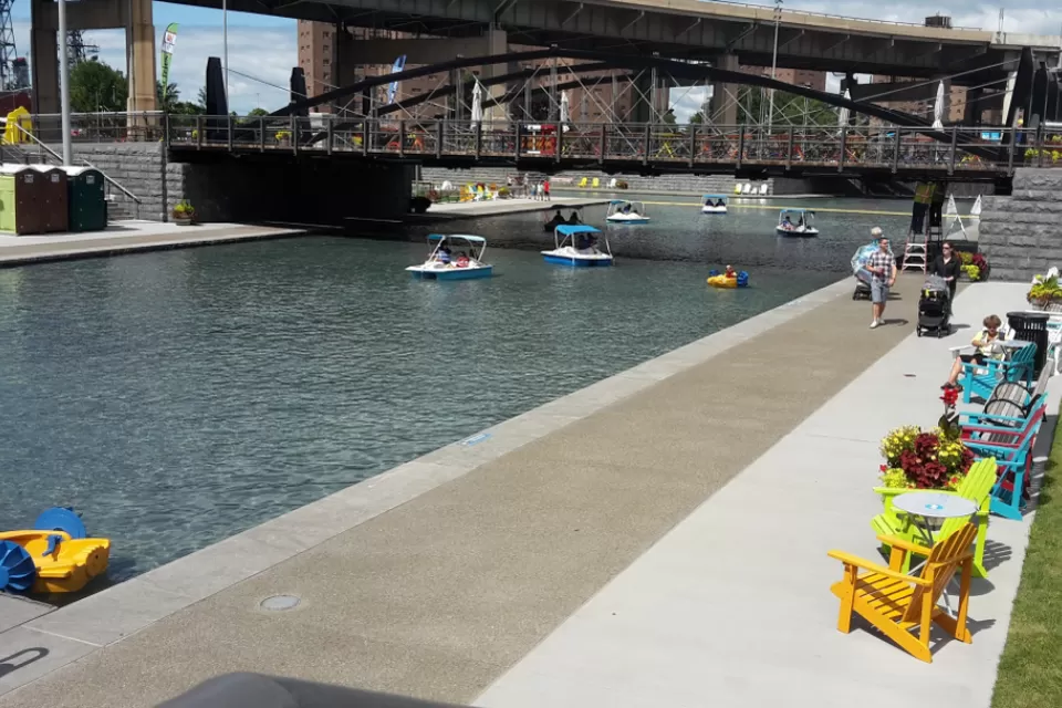 A canal under a bridge in Buffalo is lined by chairs and tables, people walking about, looking at peddle boats on the water.